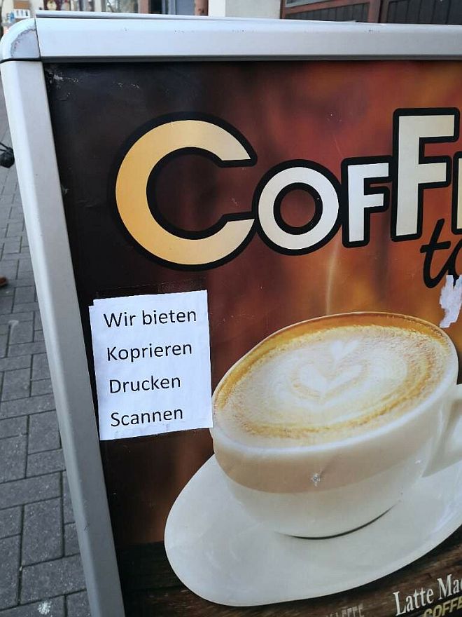 Plakat auf dem Gehweg mit Kaffeewerbung und einem Papieraufkleber "Wir bieten koprieren, drucken, scannen"