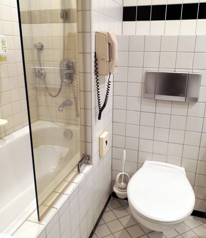 Foto zeigt ein Hotelbadezimmer mit weißen Fliesen, Badewanne mit gläserner Duschwand und Toilette. An der gefliesten Wand zwischen Dusche und direkt neben der Toilette ist ein großes Wandtelefon mit großem Hörer und typischer Telefonhörerschnur angebracht.