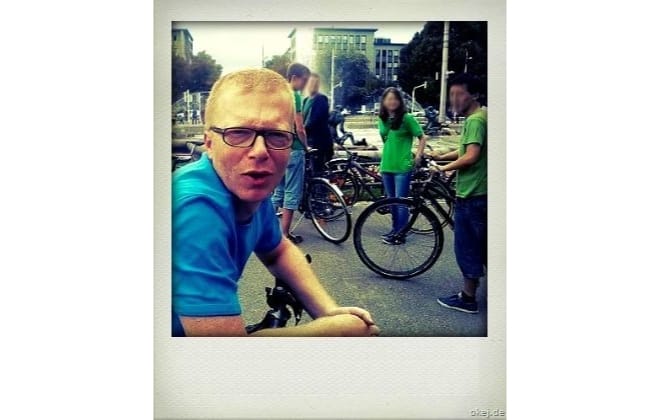 Ein Radfahrer blickt über den Lenker eines Fahrrads gebeugt in die Kamera. Im Hintergund sind andere wartende Radfahrer zu sehen.