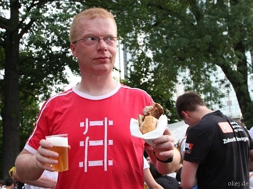 Ein Mann im rotem T-Shirt mit Grafik eines kaputten Bahngleises, in den Händen ein Plastikbecher mit Bier und Brötchen. Der Blick geht seltsam zur Seite. Im Hintergrund andere Menschen, Bäume und Hochhäuser.
