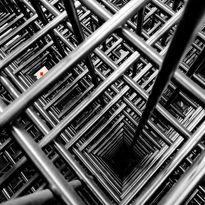Eine Vielzahl von Metallstäben kreuzen sich rechtwinklig und bilden ein regelmäßiges Geflecht in Grautönen. Eine kleine Lücke gibt den Blick auf den Hintergund frei, in der man einen Teil eines Bildes von Mondrian mit einem roten Rechteck sieht.