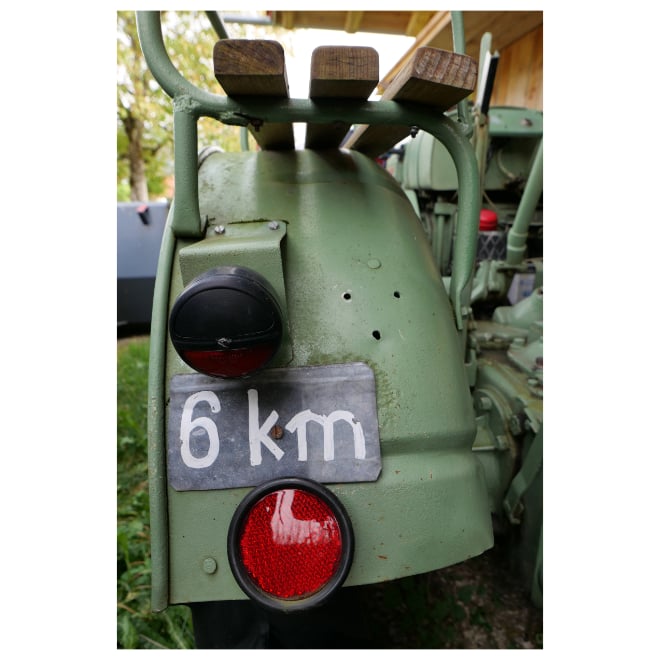 Detailaufnahme der Radabdeckung eines grünen Traktors, darauf eine Sitzgelegenheit fürs Mitfahren, zwischen Rücklicht und Reflektor ein schwarzes Schild mit aufgemalter Schrift "6 km".