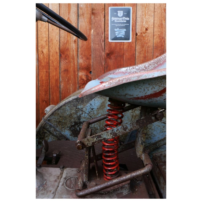 Detailaufnahme eines ehemals wohl hellblauen Traktors vor einer Holzwand mit Brauereiwerbung. Teile von Radabdeckung, Lenkrad und Sitz sind zu sehen. Die Sitzfederung mit einer rot lackierten Feder ist im Mittelpunkt.