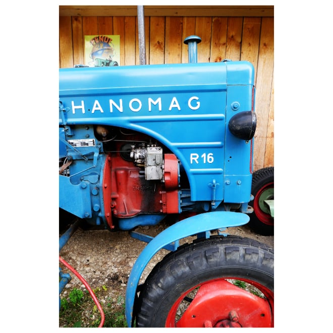 Vorderer Teil eines strahlend blauen Traktors der Marke Hanomag mit roten Details vor einer Holzwand.
