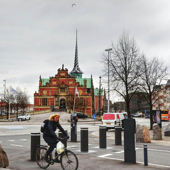 Die historische Börse in Kopenhagen mit dem markanten Turm aus ineinander gedrehten Schwänzen von Drachen und vielen Verzierungen an der Fassade ist von der Seite zu sehen. Davor eine Straßenszene, eine Radlerin fährt im Vordergund quer durchs Bild.