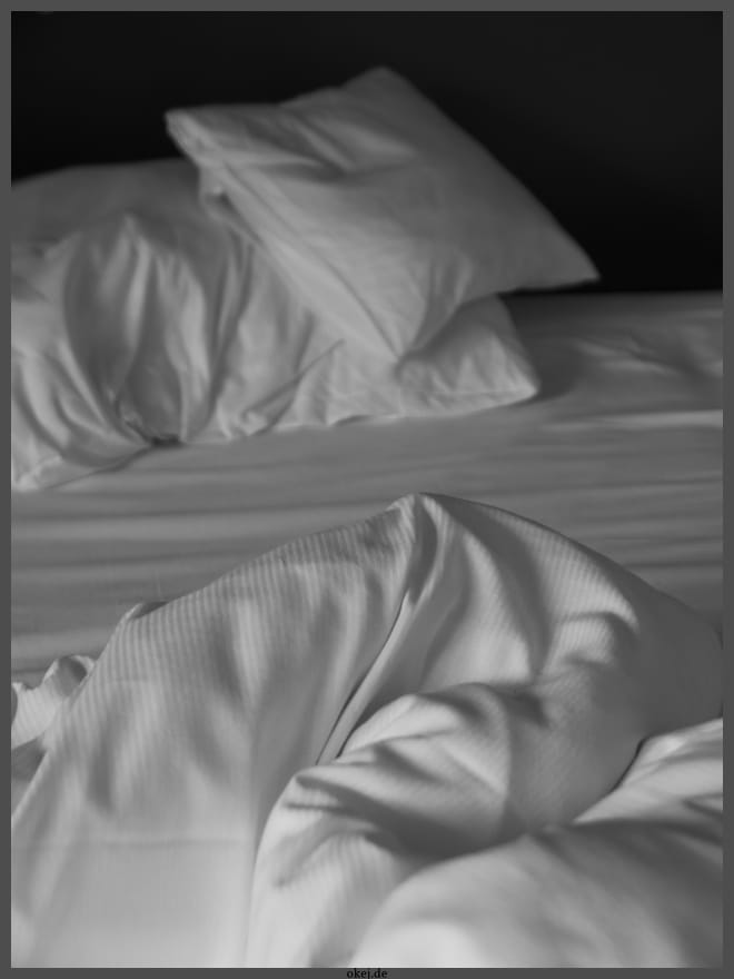 Schwarzweiß Foto von Details eines Hotelbetts. Vor schwarzer Rückwand sind zwei weiße Kissen unterschiedlicher Größe und weiße Bettwäsche zu erkennen. Aua Form und Falten kann man schließen, dass das Bett benutzt wurde.