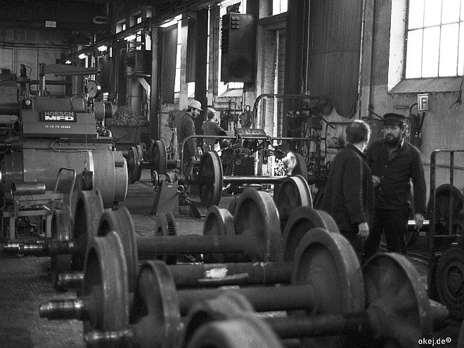 Schwarz-weiß Foto aus einer Werkstatthalle der Bahn. Ausgebaute Eisenbahnachsen im Vordergrund, im Hintergrund große Maschinen. Arbeiter in Arbeitskleidung, manche unterhalten sich.