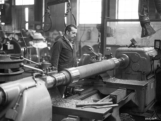 Schwarz-weiß Foto aus einer Werkstatthalle der Bahn. Ein Arbeiter beobachtet das Abdrehen einer Achse an einer großen Drehbank.