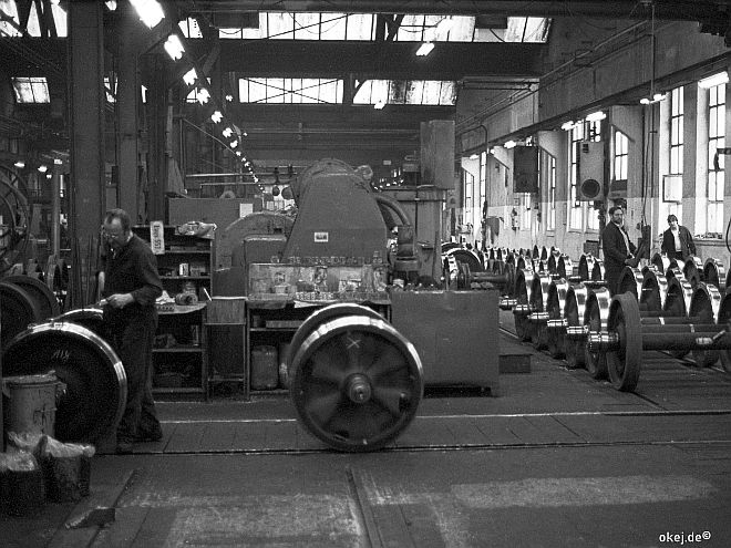 Schwarz-weiß Foto aus einer Werkstatthalle der Bahn. In Bildmitte eine große Maschine, umgeben von Eisenbahnachsen mit glänzenden Laufflächen und drei Arbeitern in schwarzer Arbeitskleidung.