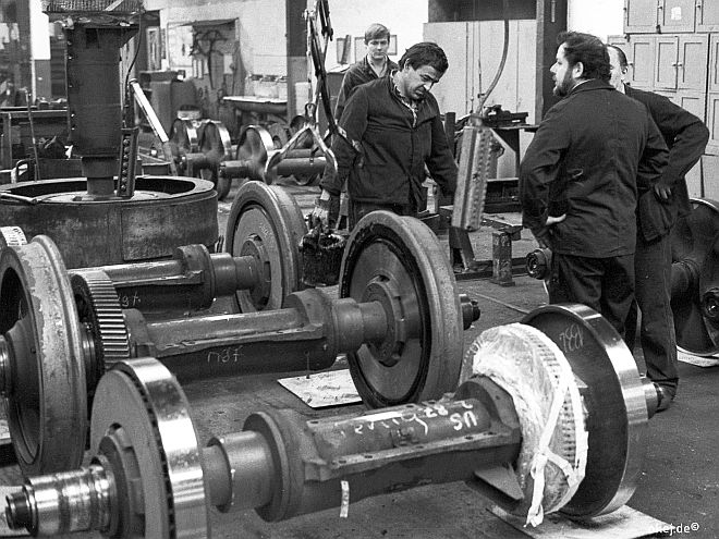 Schwarz-weiß Foto aus einer Werkstatthalle der Bahn. Es sind mehrere Achsen ohne Radkranz und zwei komplette Achsen mit Lauffläche, sowie Arbeiter in schwarzer Arbeitskleidung zu sehen.