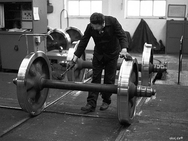 Schwarz-weiß Foto aus einer Werkstatthalle der Bahn. Eine Eisenbahnachse mit glänzenden Neuteilen wird von einem Arbeiter mit einem Pinsel behandelt.
