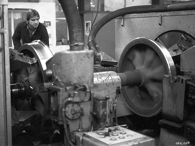Schwarz-weiß Foto aus einer Werkstatthalle der Bahn. Eine Eisenbahnachse wird in einer großen Drehbank abgedreht,. Ein Arbeiter beobachtet und steuert dies.