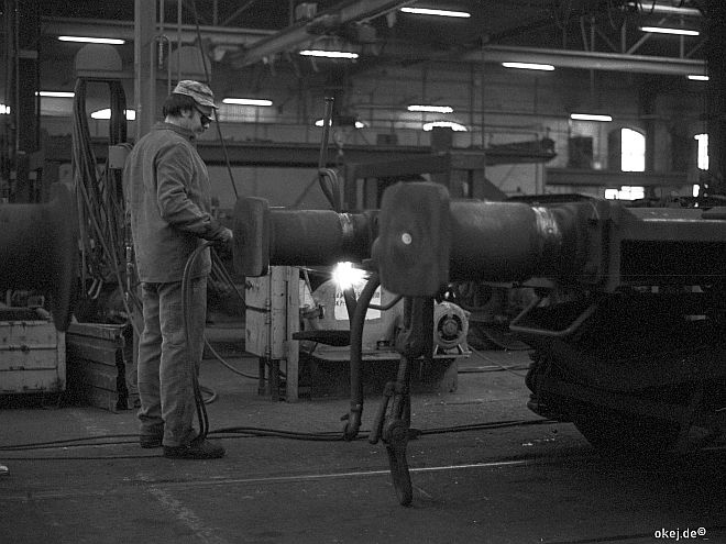 Schwarz-weiß Foto aus einer Werkstatthalle der Bahn. Ein Arbeiter mit einer Schutzbrille schweißt etwas neben den Puffern an der Stirnseite eines flachen Waggons.