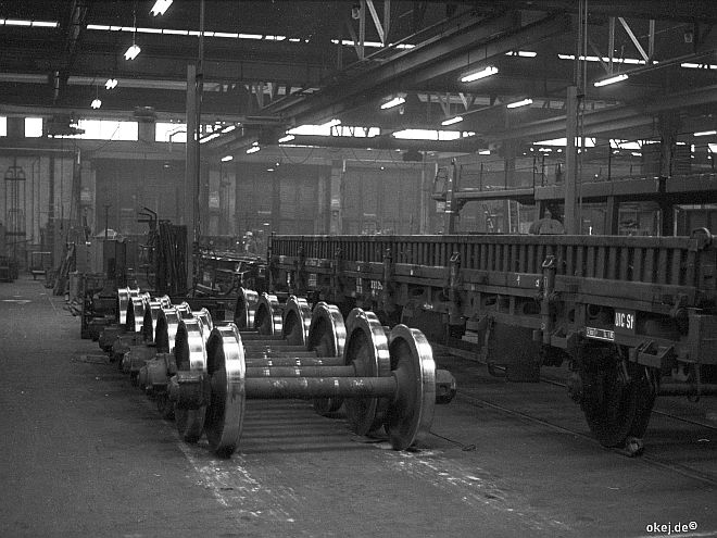 Schwarz-weiß Foto einer Werkstatthalle der Bahn. Links liegen viele blankgedrehte Achsen, rechts sind ein Flach- und ein Autotransportwagen zu sehen.