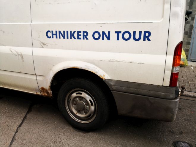 Auf dem hinteren Teil eines verrosteten, weißen und von der Seite aufgenommenen Lieferwagens steht in blauen Großbuchstaben "CHNIKER ON TOUR". Am Schmutz kann man erahnen, dass es mal "TECHNIKER" hiess.