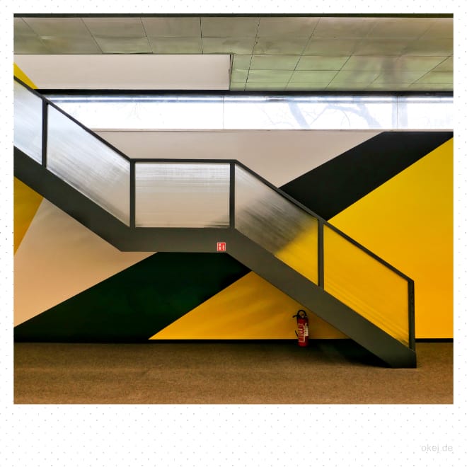 Zwischen Decke und Fußboden ist eine Wand mit Oberlicht mit großen grafischen Farbfeldern in weiß, schwarz und gelb zu sehen. Davor eine Treppe in schwarz und mit Glasverkleidungen diagonal im Bild. Darunter steht ein roter Feuerlöscher auf dem Boden.