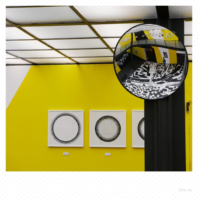 Eine gelbe Wand mit drei Bildern in schwarz-weiß. Darüber eine in Quadrate unterteilte und beleuchtete Decke Davor ein schwarzer Pfosten mit einem gekrümmten Spiegel, in dem man große Teile der Ausstellung, insbesondere den in schwarz-weiß bemalten Fußboden sieht.