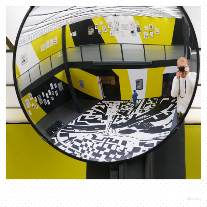 Nahaufnahme eines gekrümmten Spiegels, in dem man Teile einer Ausstellung und den in gelb, schwarz und weiß grafisch gestalteten Raum und den Fotografen sieht.