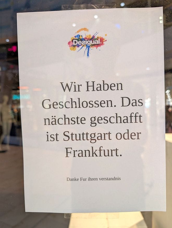 Das Bild zeigt ein Schild mit dem Desigual-Logo an einer Scheibe. Der Text darauf lautet: „Wir Haben Geschlossen. Das nächste geschafft ist Stuttgart oder Frankfurt. Danke Für ihren verständnis.“
