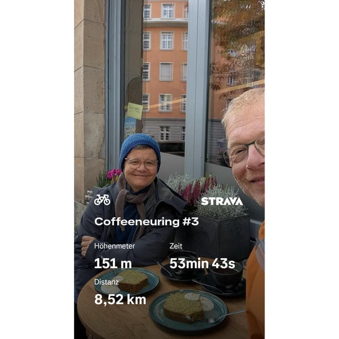 Zwei Menschen sitzen an einem kleinen runden Tisch vor einem Café und lächeln in die Kamera. Auf dem Tisch stehen zwei Tassen Kaffee und zwei Teller mit Kuchenstücken. Beide Personen tragen Brillen und warme Kleidung, eine Person eine blaue Mütze. Im Hintergrund ist die Fensterfront des Cafés mit Spiegelungen und Pflanzen zu sehen. Über das Bild sind Strava-Daten eingeblendet: Titel „Coffeeneuring #3“, 151 Meter Höhenmeter, 53 Minuten 43 Sekunden Zeit und 8,52 Kilometer Distanz.