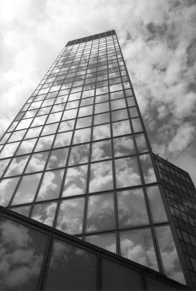 Schwarzweiß-Fotografie eines hohen, modernen Büroturms mit vollständig verglaster Fassade, in der sich dramatische Wolken am Himmel spiegeln; die Aufnahme ist stark nach oben perspektivisch verzerrt und betont die Höhe und Geometrie des Gebäudes.