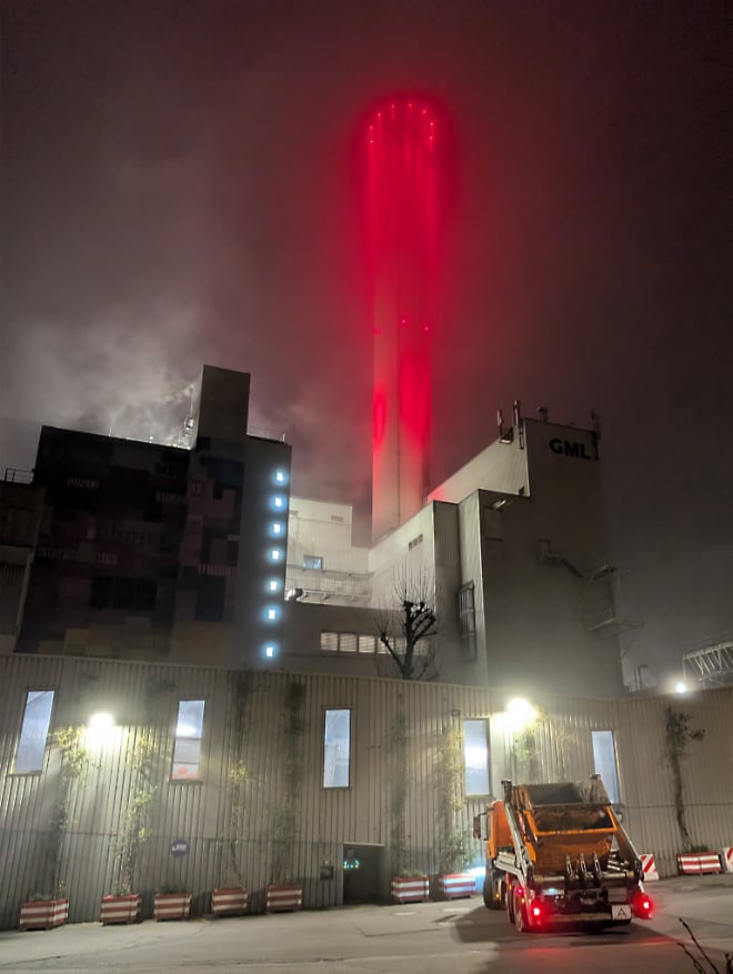Ein Müllheizkraftwerk bei Nacht, dessen hoher Schornstein in intensivem Rot von oben angestrahlt wird und oben in Dunkelheit und Nebel verschwindet. Vor der unten hell erleuchteten, industriellen Fassade steht rechts unten ein orangefarbener Müllwagen auf Asphalt.