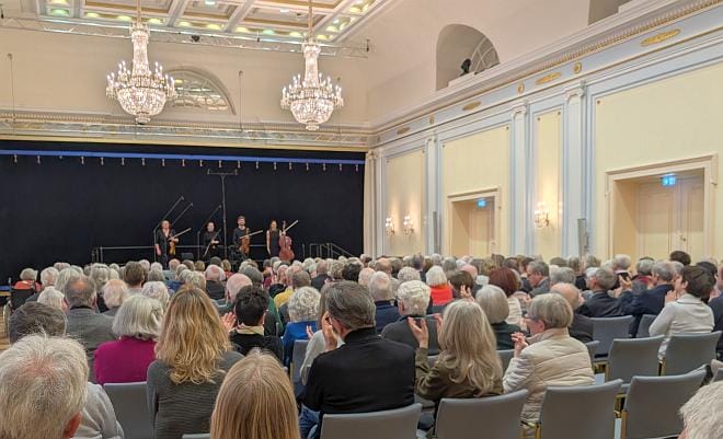 Blick in einen prunkvollen Konzertsaal mit hohen Decken und Kronleuchtern, in dem viele meist ältere Menschen in Reihen sitzen und applaudieren, während vorne auf der Bühne ein kleines Kammermusikensemble mit Streichinstrumenten den Applaus entgegennimmt.