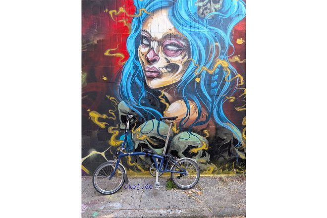 Blaues Faltfahrrad Brompton lehnt vor einer bunten Graffiti-Wand mit Porträt einer Frau mit langen blauen Haaren in Mannheim. Es sind mehrere Totenköpfe zu sehen und das Gesicht der Frau zeigt Anzeichen von Verfall.