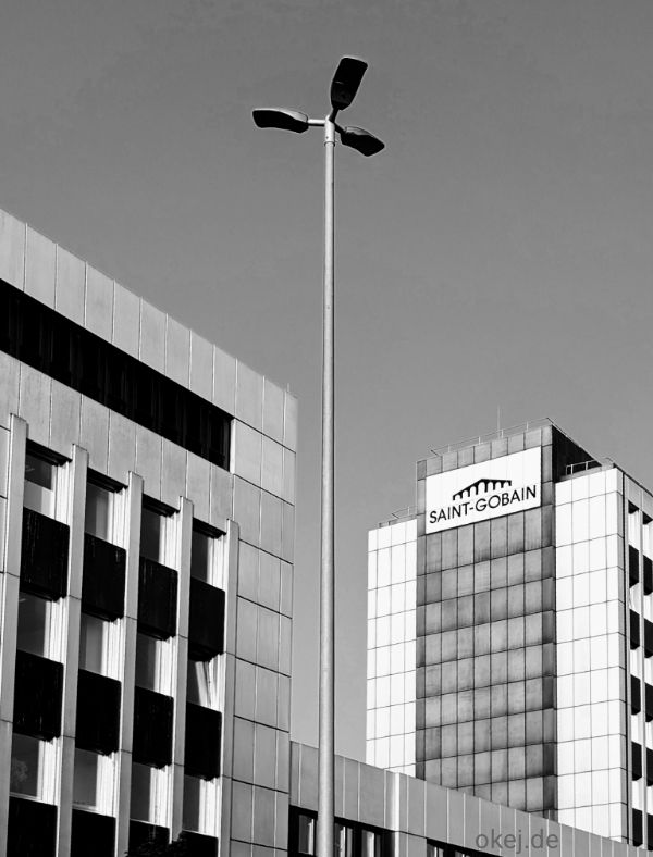 Schwarzweißfoto moderner Bürogebäude mit hoher Straßenlaterne im Vordergrund. Die drei Leuchten der Laterne sehen gegen den Himmel und in der Lücke zwischen zwei Gebäudeteilen fast wie ein Quirl aus. Auf einem Gebäude ist das Logo „Saint-Gobain“ zu sehen.